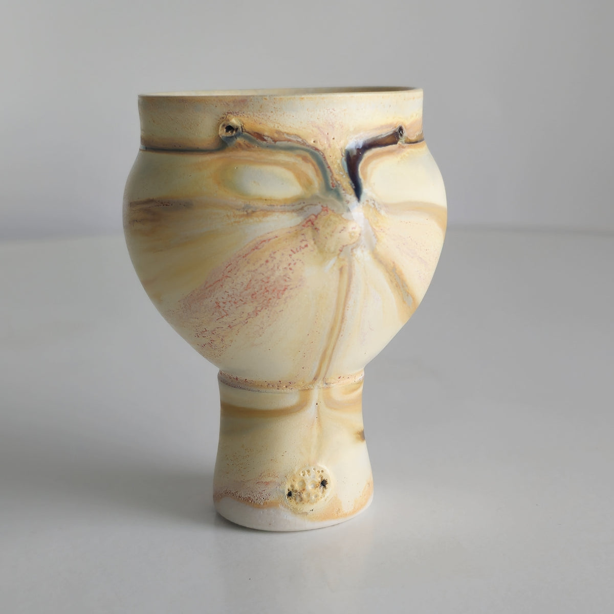 A handmade ceramic footed cup with a bulbous body and narrow base, decorated with marbled cream, beige, and light brown patterns and a small dark glaze accent near the rim, set against a plain light gray background.